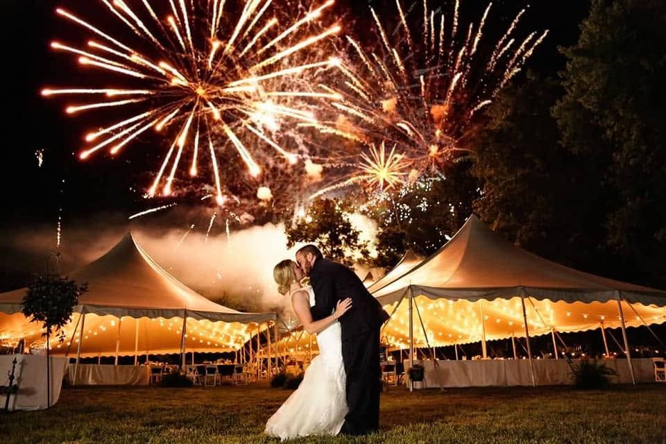Bride and groom kissing in front of lit Kink's pole tents as fireworks burst overhead
