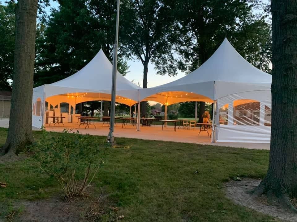 Two white high-peak tents with window sidewalls glowing with string lights at dusk