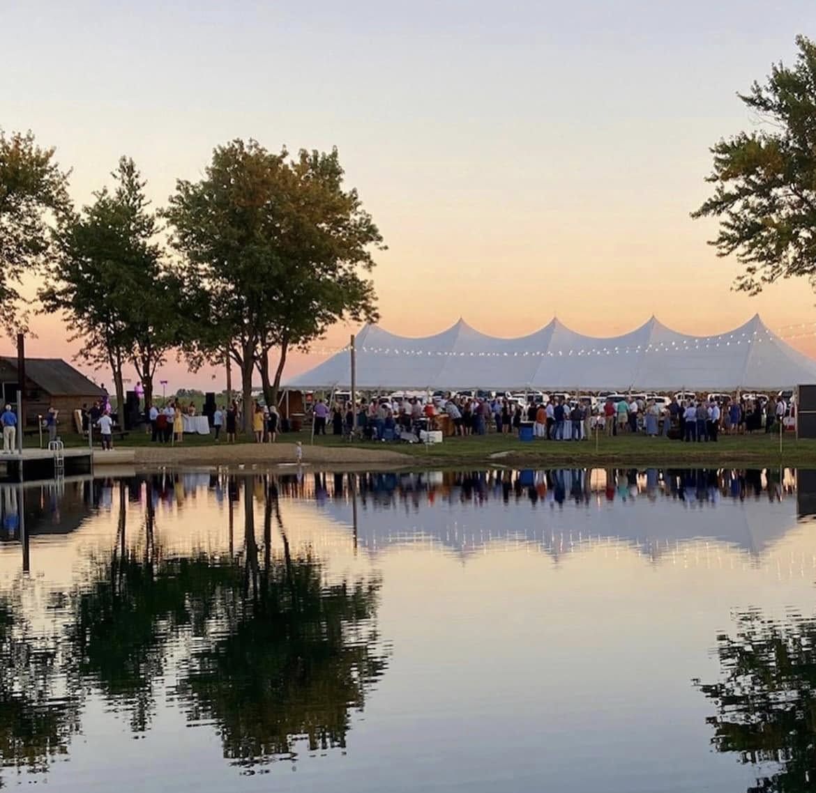 Wedding guests gathered by a pond under a high-peak tent at sunset